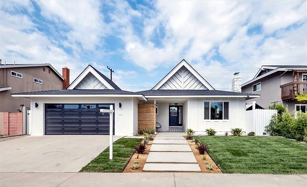 Remodeled white single-story home with board-and-batten gables, a black garage door and a paver walkway on Nyanza Drive in Huntington Beach, California